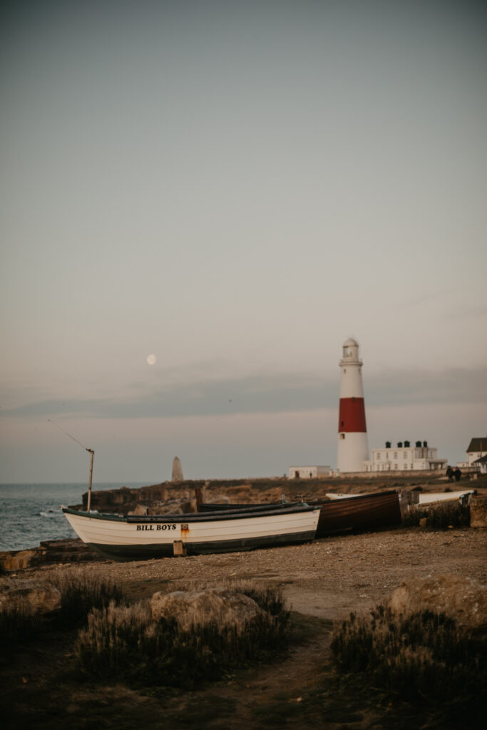 52 week photography challenge: adventure. Picture of a boat in front of Portland Bill lighthouse at sunrise, with the moon in view.