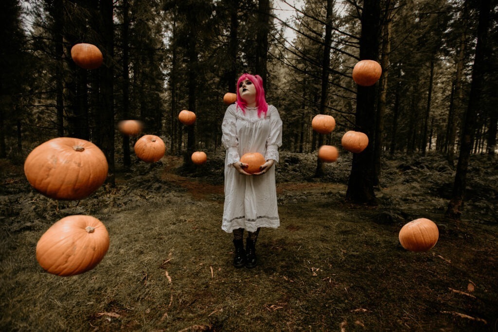Woman in white dress with pink hair, surrounded by pumpkins in the forest.