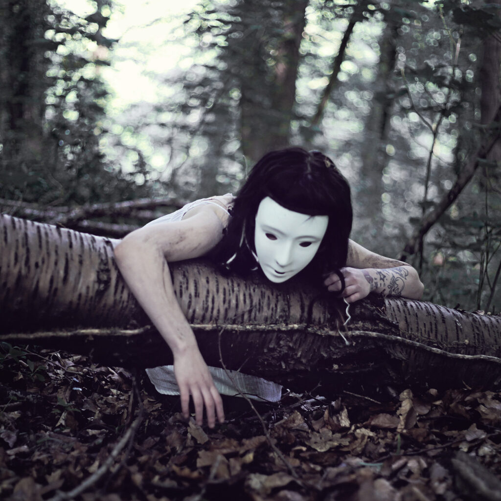 Picture of a woman in a mask laying over a fallen tree in the woods.