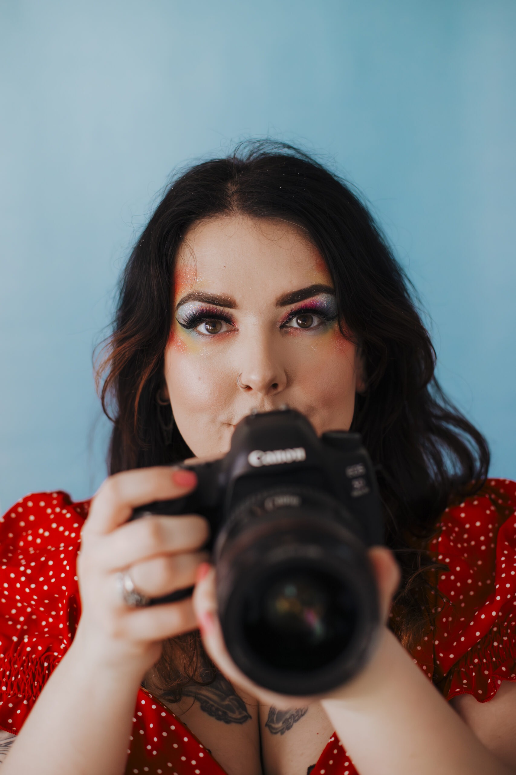 Self portrait photograph with rainbow makeup, red blouse, blue background and DSLR camera