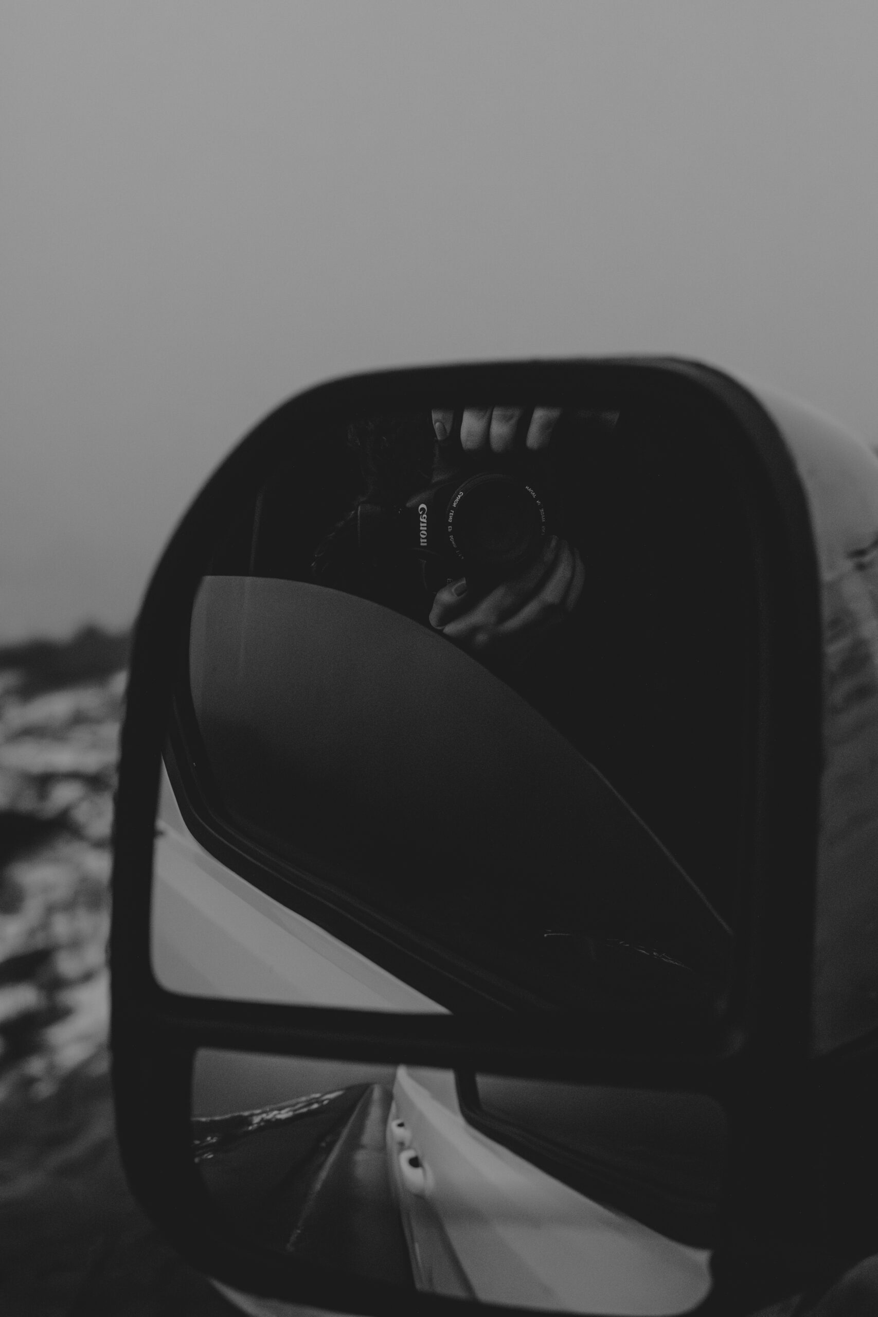 Black and white photograph of a photographer with a canon camera taking a picture of herself in a van mirror on a snowy road