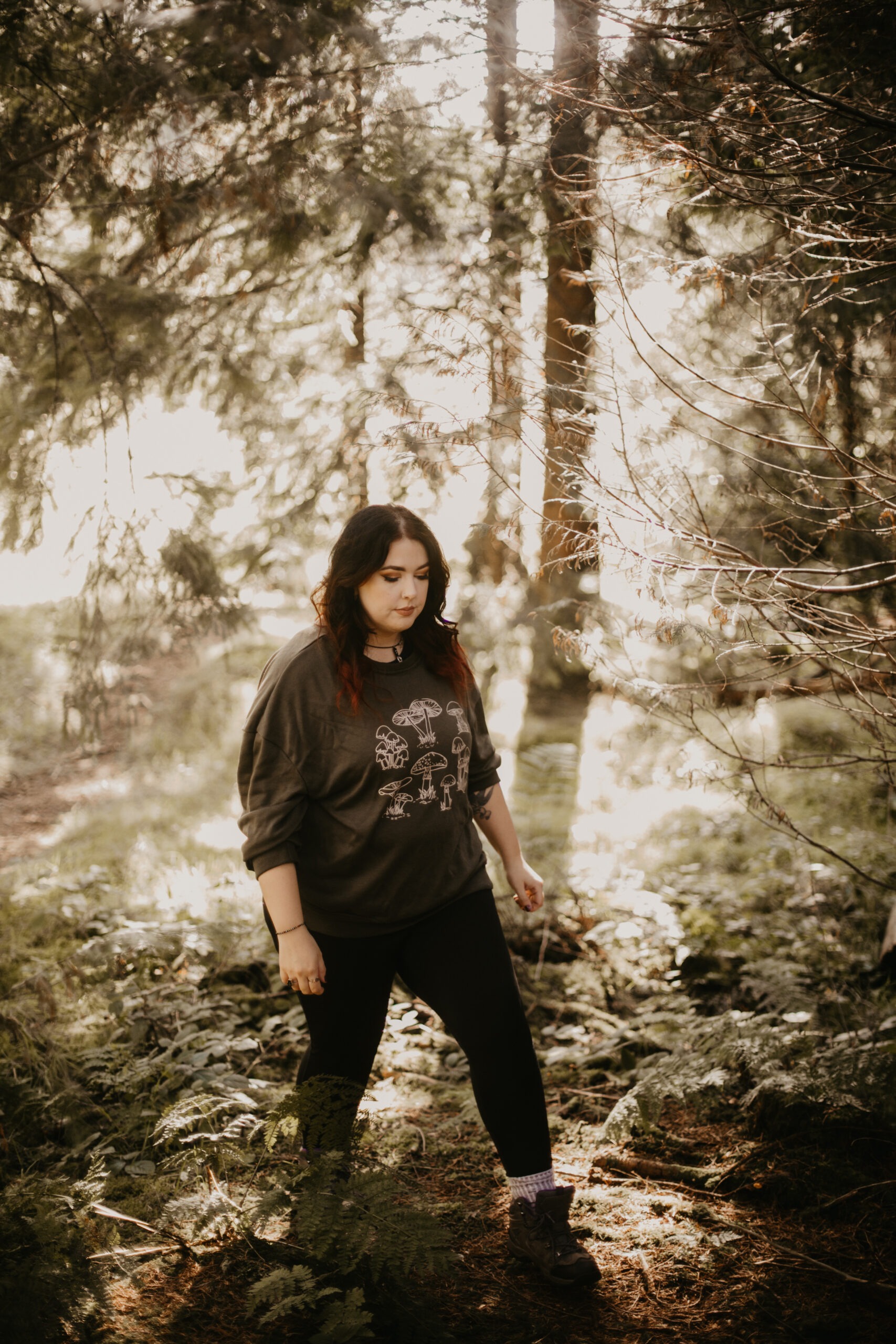 Woman walking through woods, backlit by sunlight, wearing a mushroom jumper 